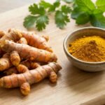 A pile of fresh turmeric roots and a bowl of golden turmeric powder on a light wooden board, with blurred green herbs.