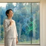 A serene woman in light clothing, hand on chest, eyes closed, stands before a window with glowing blue-green molecular structures and a blurred natural landscape.