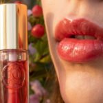 Gros plan sur une bouche aux lèvres roses brillantes à côté d'une bouteille d'huile lèvres Clarins "10 ans" sur fond floral.
