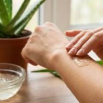 Gros plan sur une main appliquant du gel d'aloe vera sur la peau d'un bras. Une plante et un bol de gel sont visibles en arrière-plan.