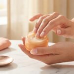 Mains appliquant la Crème Abricot Dior pour ongles. Un pot de crème est ouvert sur une table en marbre, avec une serviette rose.