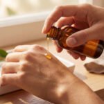 Une main verse de l'huile de nigelle d'une bouteille ambrée sur l'autre main, avec des graines de nigelle sur un plateau.
