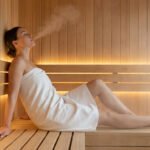 Femme détendue sur un banc de sauna en bois, yeux fermés, entourée de vapeur. Lumières chaudes en arrière-plan.