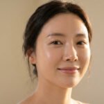 Portrait lumineux d'une femme au sourire doux, peau éclatante de santé et hydratée. Cheveux foncés attachés.