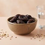 Un bol de pruneaux, un verre d'eau, des flocons d'avoine et des graines de lin sur une surface beige.