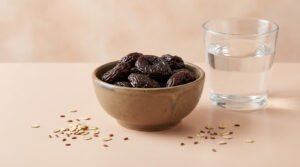 Un bol de pruneaux, un verre d'eau, des flocons d'avoine et des graines de lin sur une surface beige.