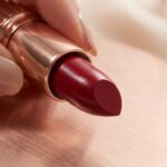 Gros plan sur un rouge à lèvres Lancôme Absolu Rouge satiné bordeaux, tenu par une main. Étui or rose.