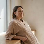 Femme mature aux cheveux bruns, chemise beige, reposant son bras sur un comptoir en bois, regardant pensivement une fenêtre ensoleillée.