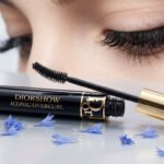 Un tube de mascara Diorshow Iconic Overcurl noir et or ouvert, sa brosse près d'un œil aux cils longs et courbés. Des pétales bleus entourent le produit.