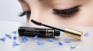 Un tube de mascara Diorshow Iconic Overcurl noir et or ouvert, sa brosse près d'un œil aux cils longs et courbés. Des pétales bleus entourent le produit.